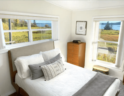Cozy bedroom with a bed, decorative pillows, and large windows showcasing a scenic view of nature outside.