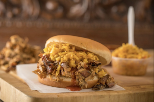A pulled pork sandwich topped with yellow rice, served with a side of rice in a cup, on a wooden surface.