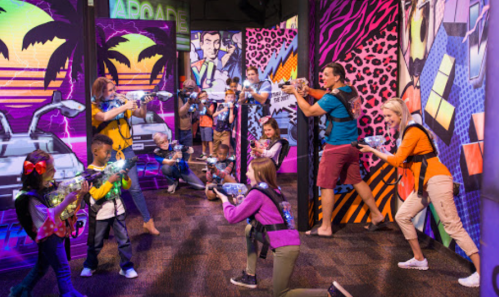 A group of children and adults play laser tag in a colorful, neon-themed arcade setting.