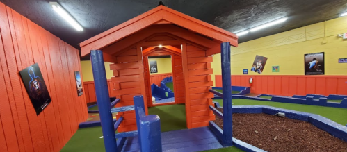 Colorful indoor mini-golf course featuring a red playhouse and blue pathways surrounded by green turf.