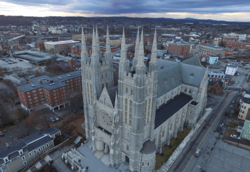 Aerial view of a large, ornate cathedral with spires, surrounded by a cityscape and mountains in the background.