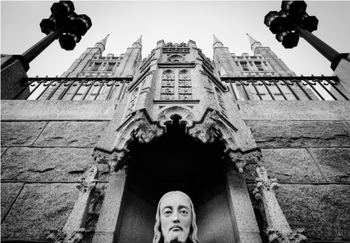 A close-up of a statue of Christ at the base of a grand, gothic-style building with spires. Black and white image.