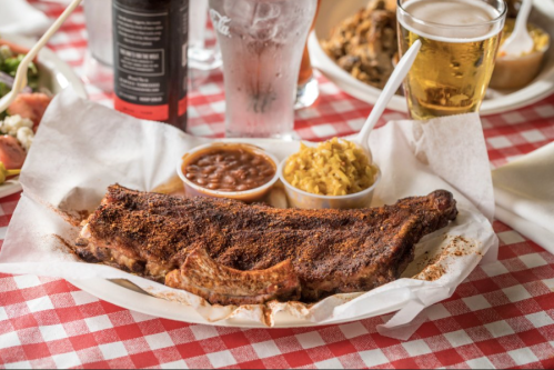 A plate of seasoned ribs with sides of baked beans and macaroni, accompanied by a glass of beer on a checkered tablecloth.