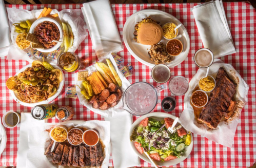 A table spread with various barbecue dishes, sides, drinks, and a salad on a red and white checkered tablecloth.
