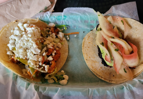 Two tacos on a green paper wrapper: one with meat, cheese, and vegetables; the other with black beans and pickled vegetables.