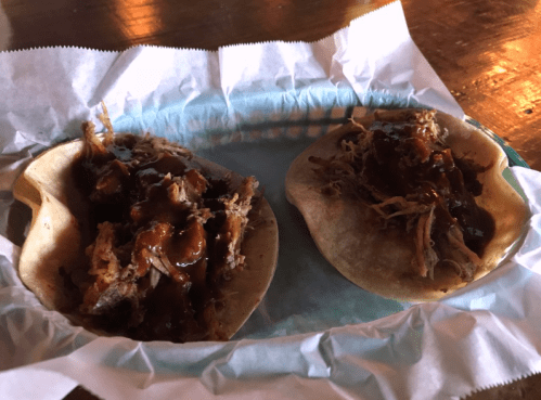 Two soft tacos filled with shredded meat and drizzled with barbecue sauce, served on a paper-lined tray.
