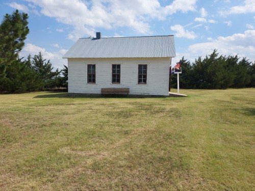 A small white house with a metal roof, surrounded by green grass and trees under a partly cloudy sky.