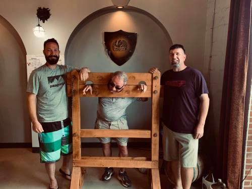 Two men stand beside a wooden stocks device, with a third man playfully locked inside, all smiling in a cozy room.