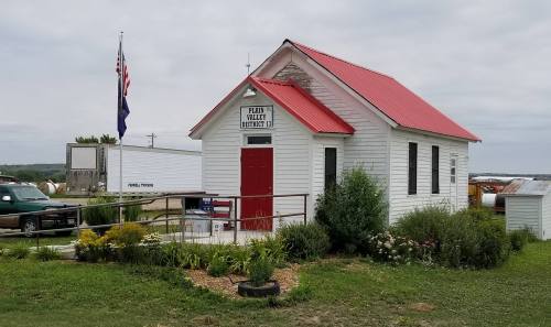 A small white building with a red roof, labeled "Plain Valley District 13," surrounded by greenery and an American flag.
