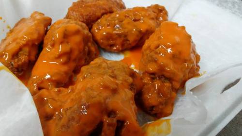 A close-up of crispy chicken wings coated in a thick, spicy orange sauce on a white paper-lined tray.