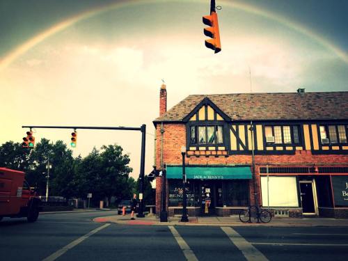 A quaint brick building with a rainbow arching overhead, near a traffic light and a parked bicycle.