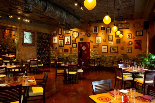 Cozy restaurant interior with wooden tables, chairs, and colorful wall art, illuminated by yellow pendant lights.