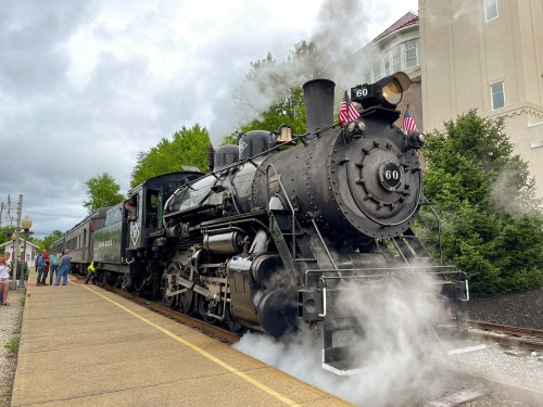 A vintage steam locomotive with smoke billowing, parked at a station with people nearby and a cloudy sky.