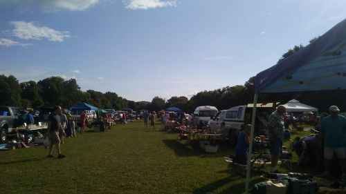 A bustling outdoor market with tents, vendors, and shoppers on a sunny day, surrounded by green fields and trees.