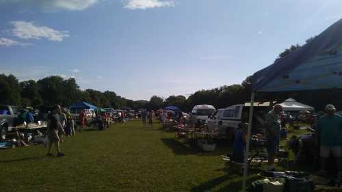 A bustling outdoor market with tents, vendors, and shoppers on a sunny day, surrounded by green fields and trees.