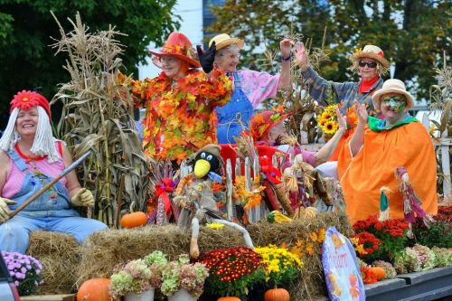 A colorful parade float with cheerful participants in autumn costumes, surrounded by hay, pumpkins, and fall decorations.