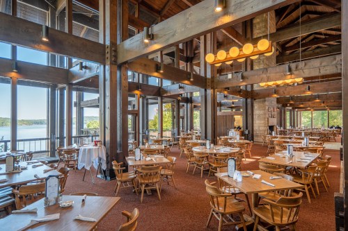 Spacious restaurant with wooden beams, large windows, and tables set for dining, overlooking a scenic lake view.