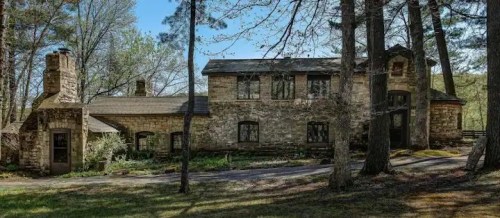 A rustic stone house surrounded by trees, featuring a chimney and large windows, set in a natural landscape.