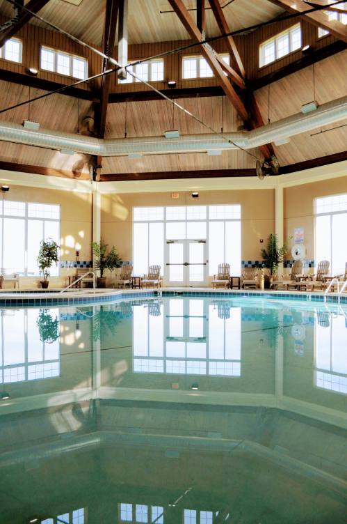 Indoor pool area with a high wooden ceiling, large windows, and lounge chairs around the water.