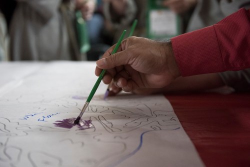 A hand holding a green paintbrush applies purple paint to a large paper with outlines and handwritten notes.