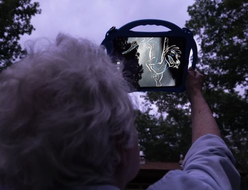 A person holds a tablet displaying a drawing of a horse against a dark, cloudy sky. Trees are visible in the background.