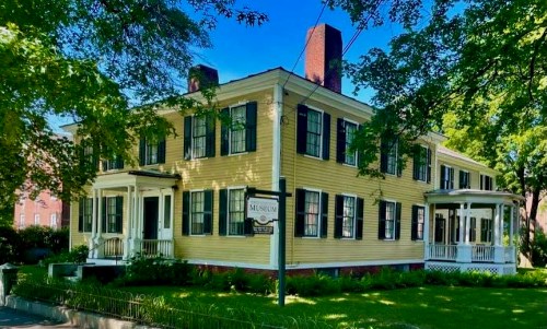 A large yellow historic house with black shutters, featuring a porch and a sign indicating it is a museum.
