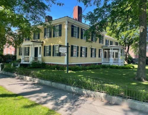 A large yellow house with black shutters, surrounded by greenery and a sidewalk, under a clear blue sky.