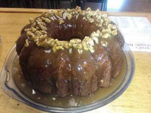 A round bundt cake topped with a glossy caramel glaze and chopped walnuts, sitting on a clear glass plate.