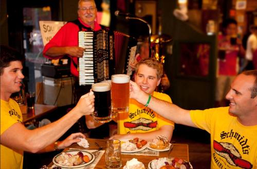 Three friends in yellow shirts toast with large beers at a festive table, while an accordion player performs in the background.