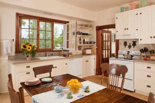 Bright kitchen with a wooden table, sunflowers, and a window overlooking greenery. Plates and lemons are on the table.