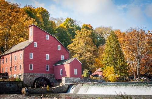 A red historic mill beside a flowing river, surrounded by vibrant autumn foliage and a gentle waterfall.