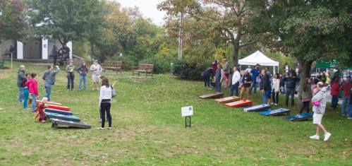 A lively outdoor event with people playing cornhole on a grassy field, surrounded by trees and spectators.