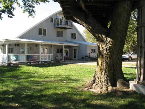 A large house with a triangular roof, porch, and a tree in the foreground, set in a grassy area.