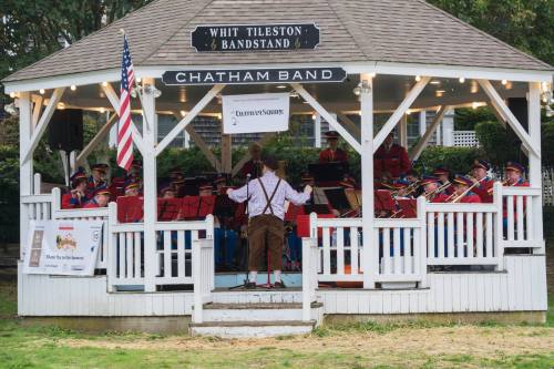 A band performs in a gazebo, with a conductor leading the musicians in colorful uniforms. An American flag is visible.