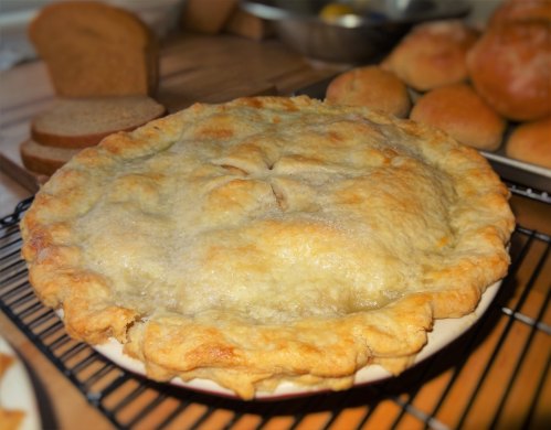 A freshly baked pie with a golden crust, resting on a cooling rack, surrounded by rolls and slices of bread.