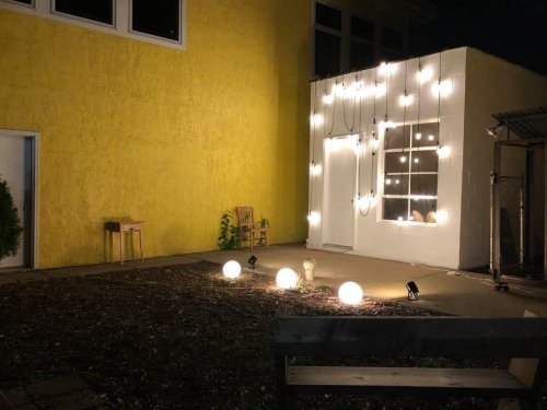 A cozy outdoor space at night, featuring a yellow wall, a lit-up white building, and glowing orbs on the ground.
