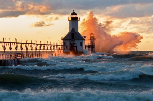 A lighthouse stands on a pier as waves crash dramatically against it during a vibrant sunset.