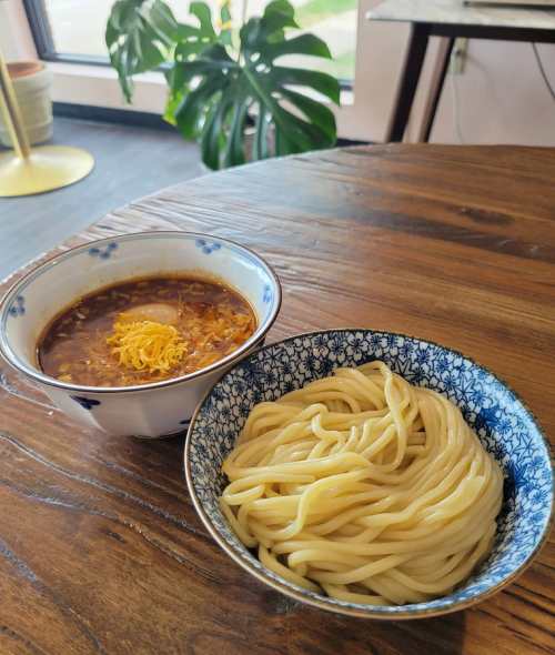 A bowl of noodles next to a bowl of rich, dark broth topped with cheese and an egg, on a wooden table.