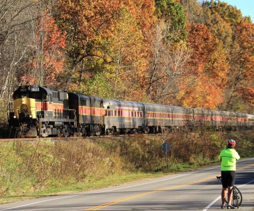 A cyclist watches a vintage train pass by on a scenic road lined with autumn trees.