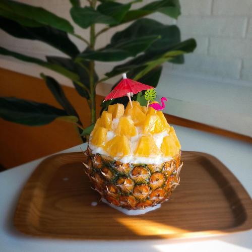A pineapple filled with cream and topped with pineapple chunks, a small umbrella, and a flamingo decoration on a wooden plate.