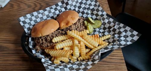 Two sandwich buns filled with seasoned ground meat, served with crinkle-cut fries and pickles on a checkered paper plate.