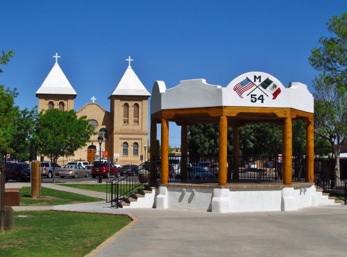 Historic church towers behind a gazebo in a park, with flags representing the U.S. and Mexico. Clear blue sky above.