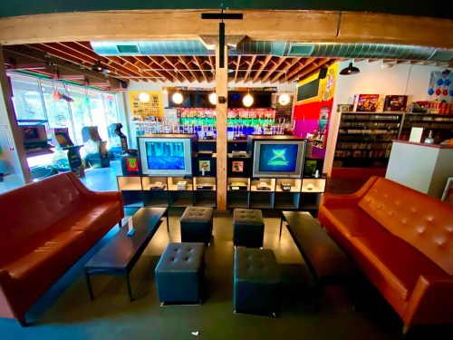 A cozy lounge area with two sofas, coffee tables, and retro TVs, surrounded by arcade games and a bar in the background.