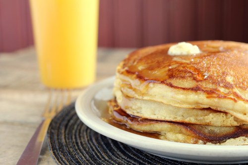 A stack of fluffy pancakes topped with butter and syrup, next to a glass of orange juice on a table.