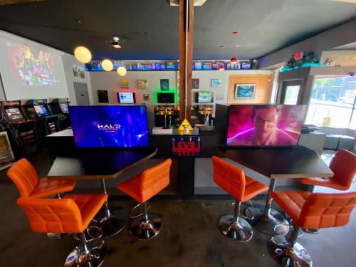 A gaming lounge with multiple screens, arcade machines, and orange bar stools arranged around a central table.
