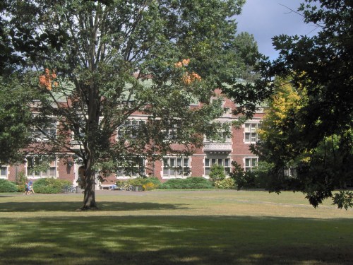 A brick building surrounded by trees and greenery, with people walking on a path in a sunny park setting.