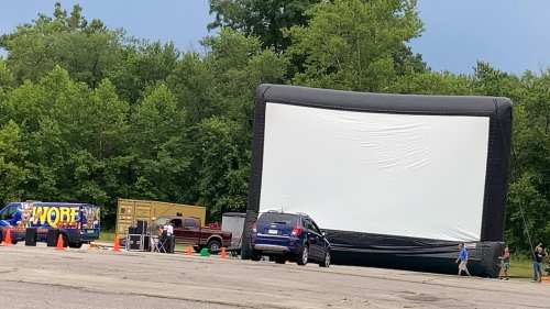 A large inflatable movie screen set up outdoors, with cars parked nearby and people walking around in a green area.
