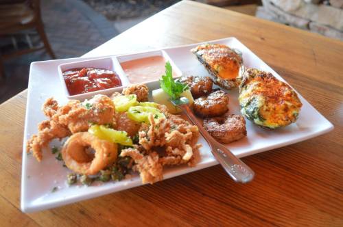 A white plate with fried calamari, shrimp, stuffed oysters, and dipping sauces on a wooden table.