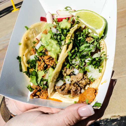 A hand holding two tacos filled with meat, topped with cilantro, onions, and lime, served in a white container.