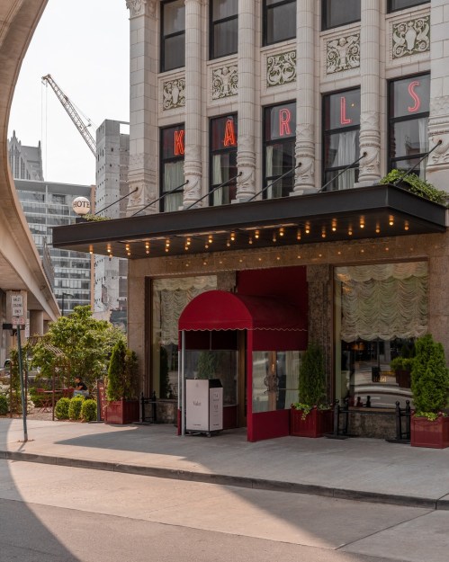 Entrance of a building with "KARLS" sign, red awning, and decorative plants, set in an urban environment.